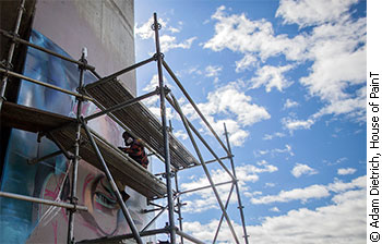 A graffiti artist is sitting on a high scaffold and painting on one side of the Dunbar Bridge