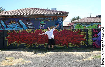 With his arms far apart, Mike Gall is posing in front of his graffiti piece that reads "Janice" in red.