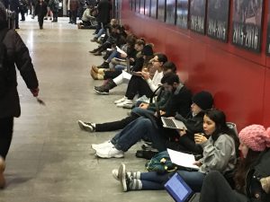 Students sitting in the hall outside the exam room with their computers open